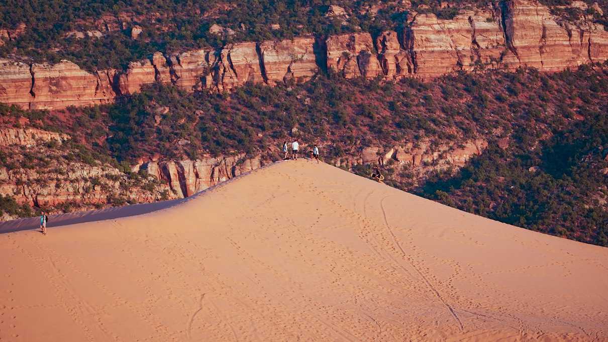 Several hikers on top of a sunlit pink sand dune with striped cliff face in background