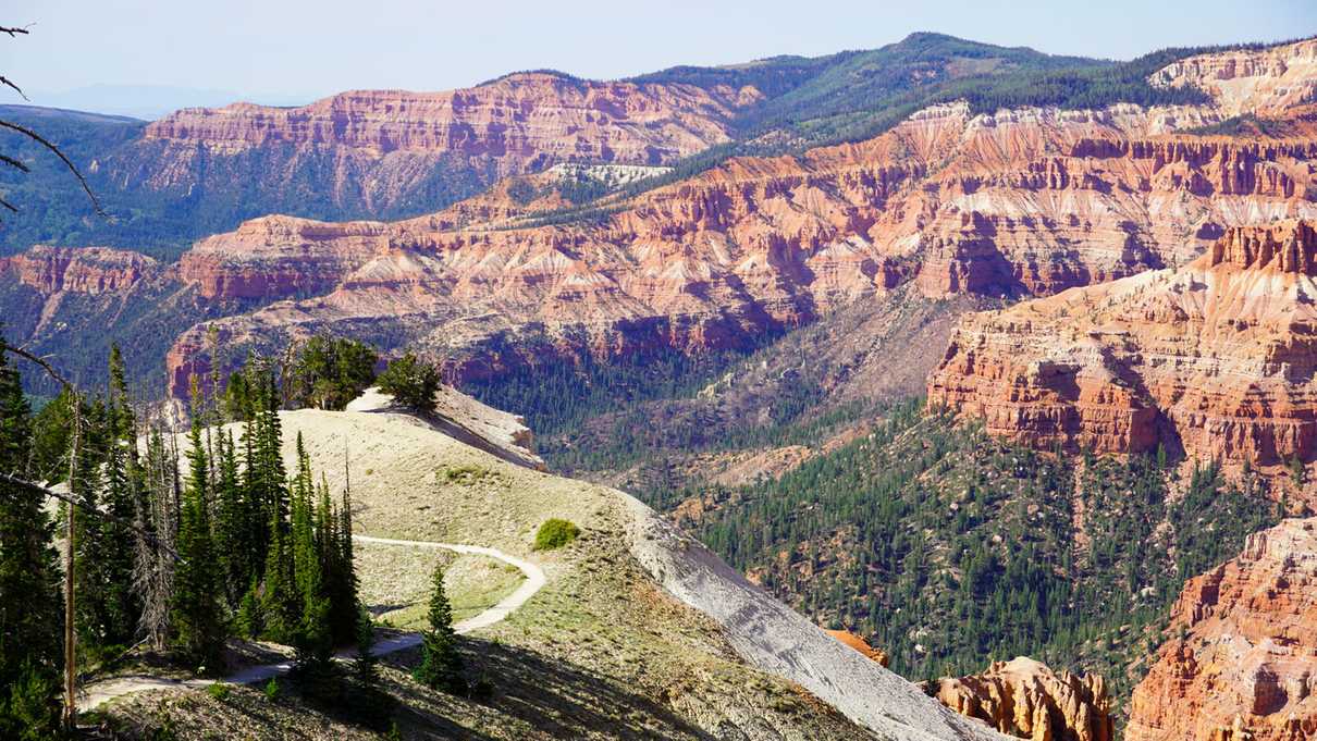 Trail wanders through grassy overlook with views of a canyon featuring bands of colored rock below