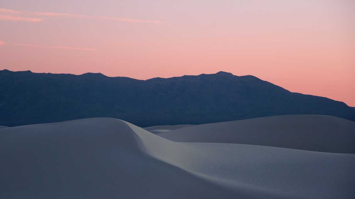 White sand dune in foreground with mountains in background against a pink sky