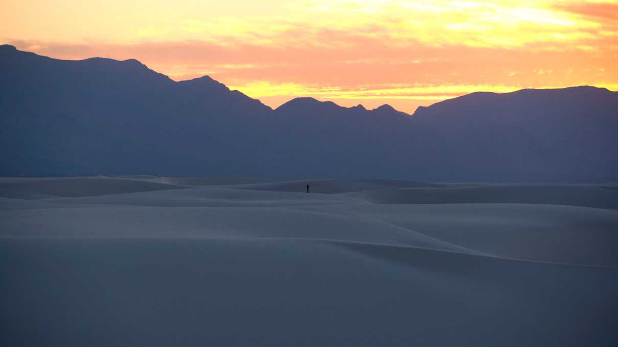 Distant figure alone on dunefield with mountains in background and orange sunset